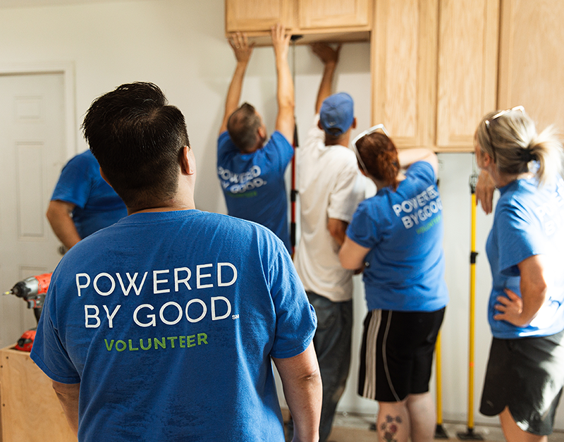 A man wearing a shirt that says "Powered by Good Volunteer" helps five team members hang cabinets