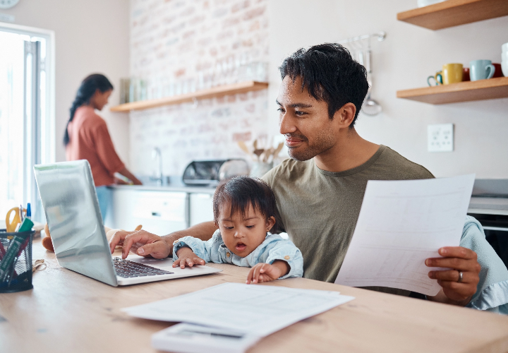 Father working on laptop with son on his lap, sitting at the kitchen table. 