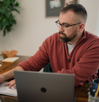  A man with glasses sits at a table with a silver laptop and writing in a notebook