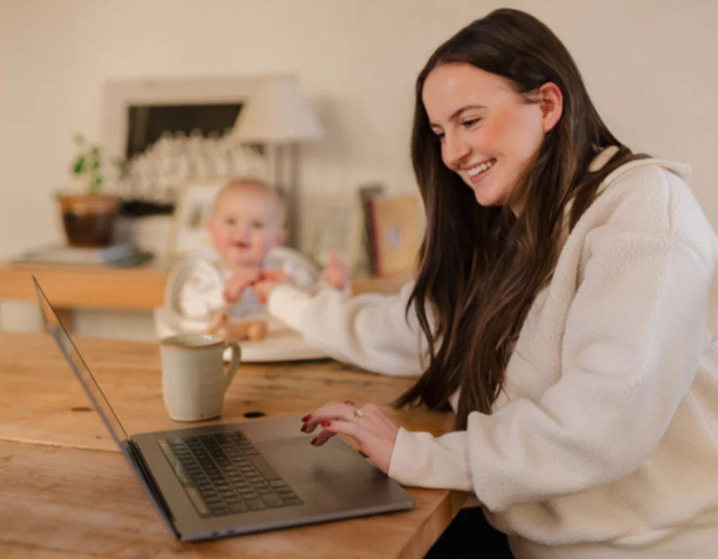 A woman types on a laptop at a kitchen table while feeding a baby