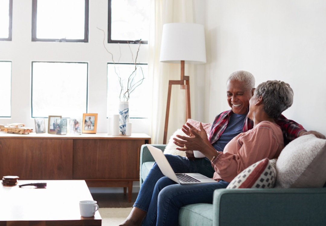 An older couple cit on a couch and laugh behind a laptop 