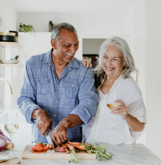 In a kitchen, an older man cuts vegetables while an older woman laughs and holds a glass of wine