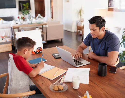 A man types on a laptop at a kitchen table while a young boy across from him writes in a notebook 