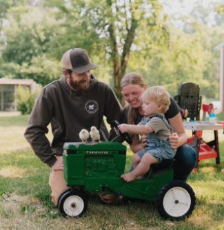 Young couple on farm, with their toddler son riding on mini tractor