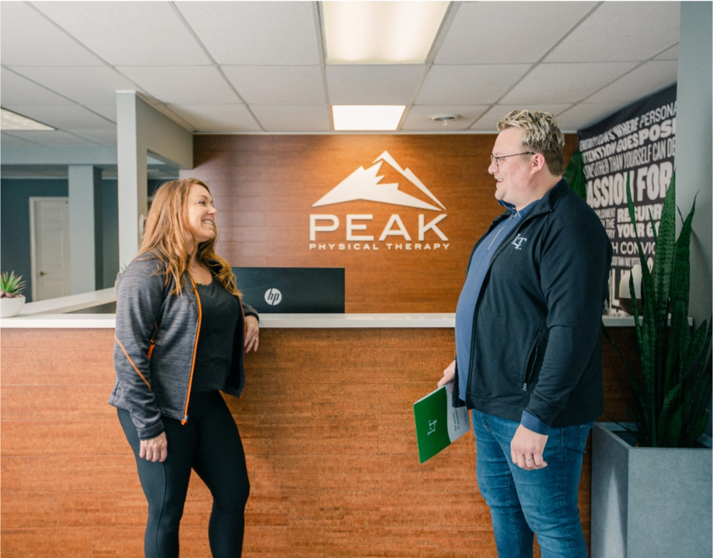 Amy, the owner of Peak Physical Therapy, stands at the front desk of her office and speaks to a Lake Trust team member