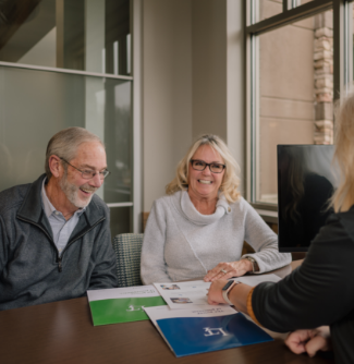 An older couple smile and laugh at a desk in a Lake Trust branch as a team member points to a piece of paper