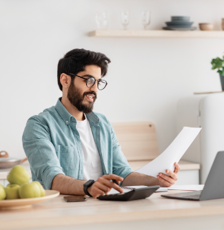 A man with a beard and glasses sits at a table and looks at a piece of paper while typing in a calculator