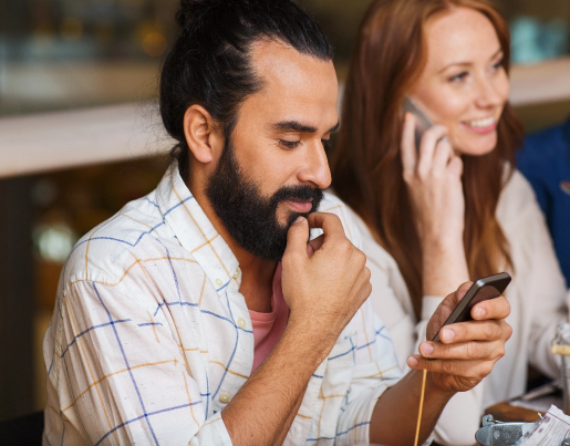 A man with a thick beard texts on his phone while a woman in the background talks on a phone