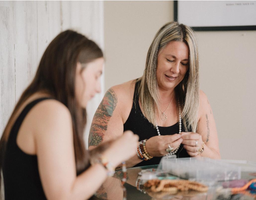 Nicole, the owner of More Love Collective, sits with another woman while making a bracelet