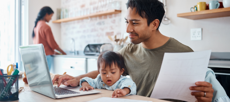 Father working on laptop with son on his lap, sitting at the kitchen table. 