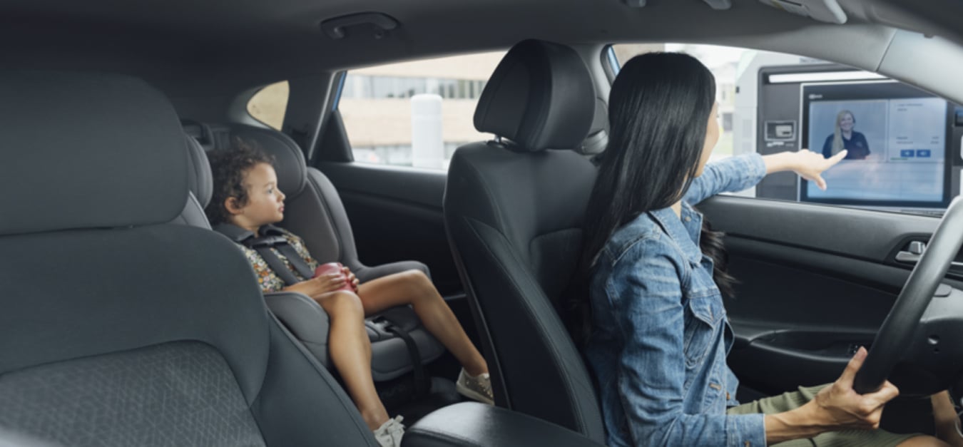 A woman visits a Lake Trust video teller machine from her car and a toddler is in a car seat in the back of the car