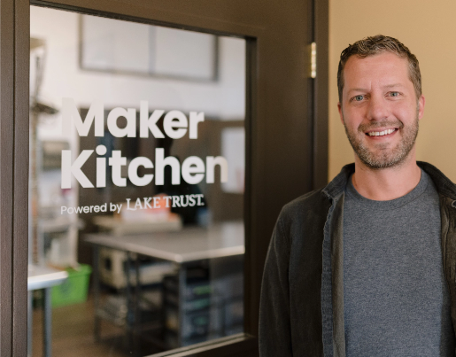 A man stands in front of a sign that says "Maker Kitchen Powered by Lake Trust"