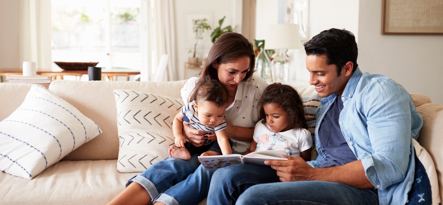 Two parents sit on a couch with a baby and toddler and read a book