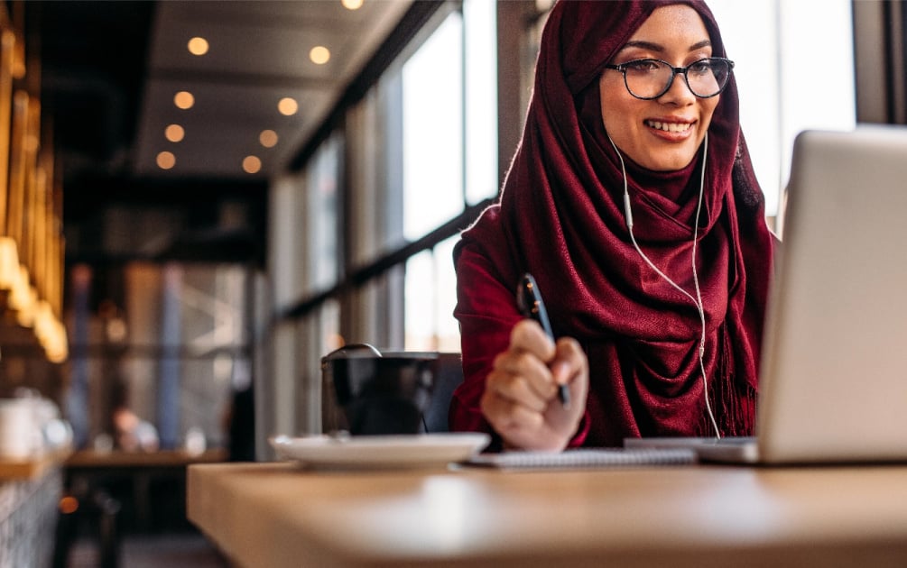 A woman with glasses sits in front of a laptop and a cup of coffee sits next to her