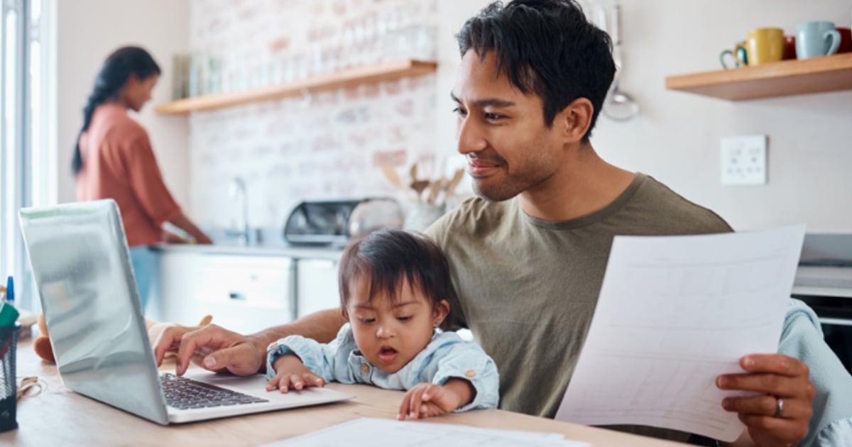 Man usiing a laptop at the kitchen table with papers in his hand and a young child on his lap.