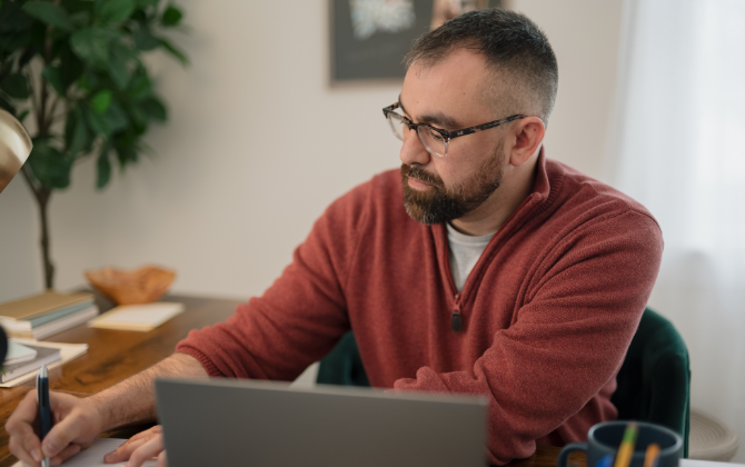  A man with glasses sits at a table with a silver laptop and writing in a notebook