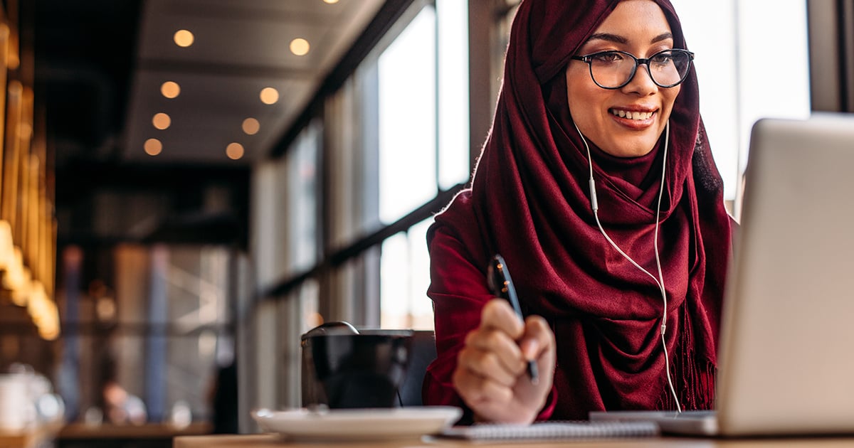 A woman with glasses sits in front of a laptop and a cup of coffee sits next to her