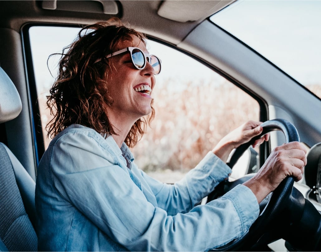 A woman in sunglasses sits in a car with her hands on the steering wheel