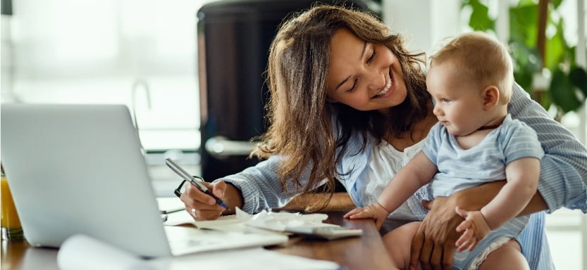 A woman holds a baby while paying bills