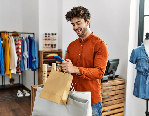 A man holds three shopping bags in a clothing store and texts on his phone