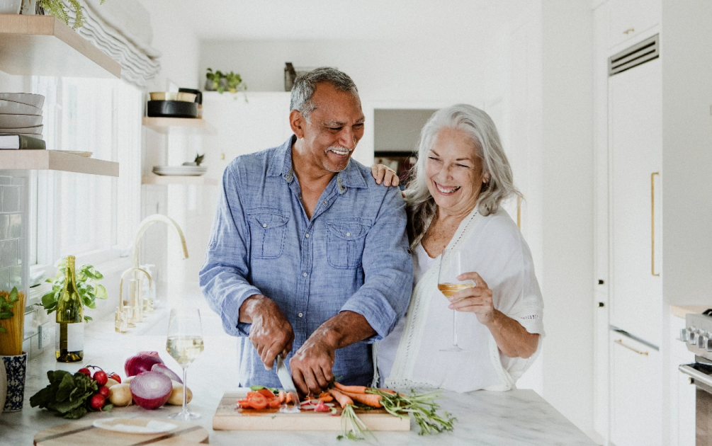 In a kitchen, an older man cuts vegetables while an older woman laughs and holds a glass of wine