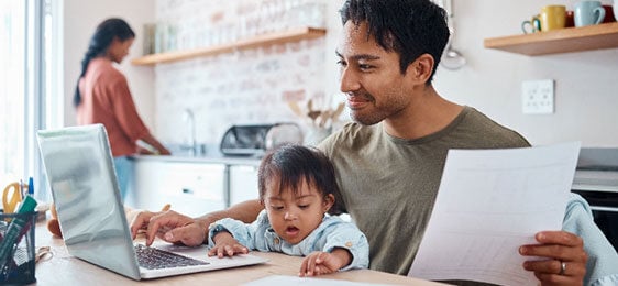 Father sitting at kitchen table using laptop with baby on lap.