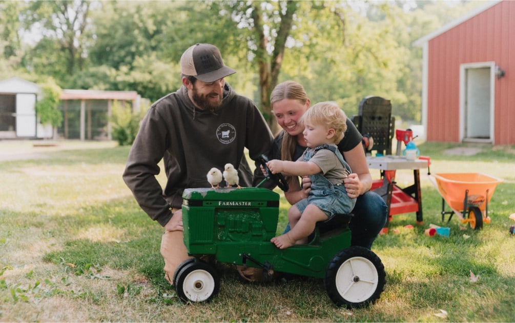 Young couple on farm, with their toddler son riding on mini tractor. 