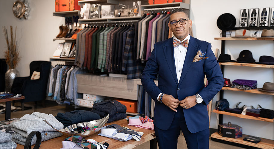 Ne'Gyle, the owner of Bleu Bowtique, is wearing a blue suit and stands in front of clothing for sale in his store