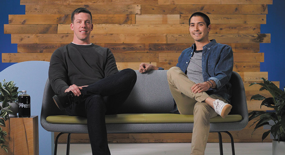 Kevin and Nick, the owners of Tapped Coffee, sit on a gray and green couch in front of a wood wall