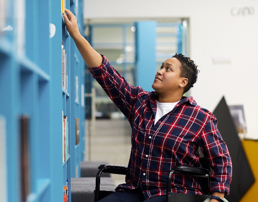 A person in a wheelchair is in a library and reaches up to take a book off of a shelf