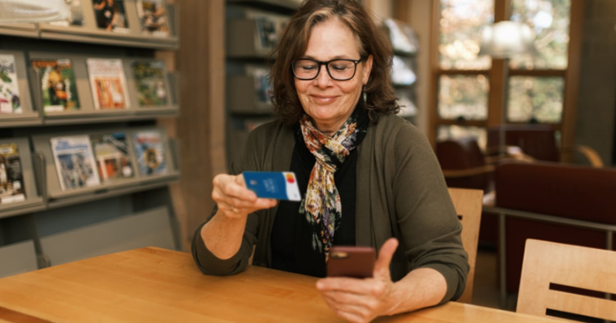 An older woman in glasses looks at her phone and holds a Lake Trust debit card