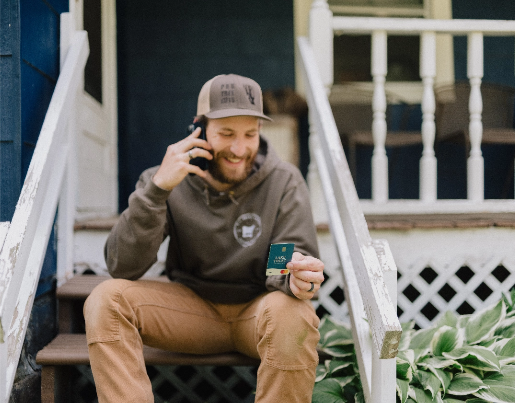 A man in a baseball hat sits on porch stairs while talking into a cell phone and holding a Lake Trust debit card