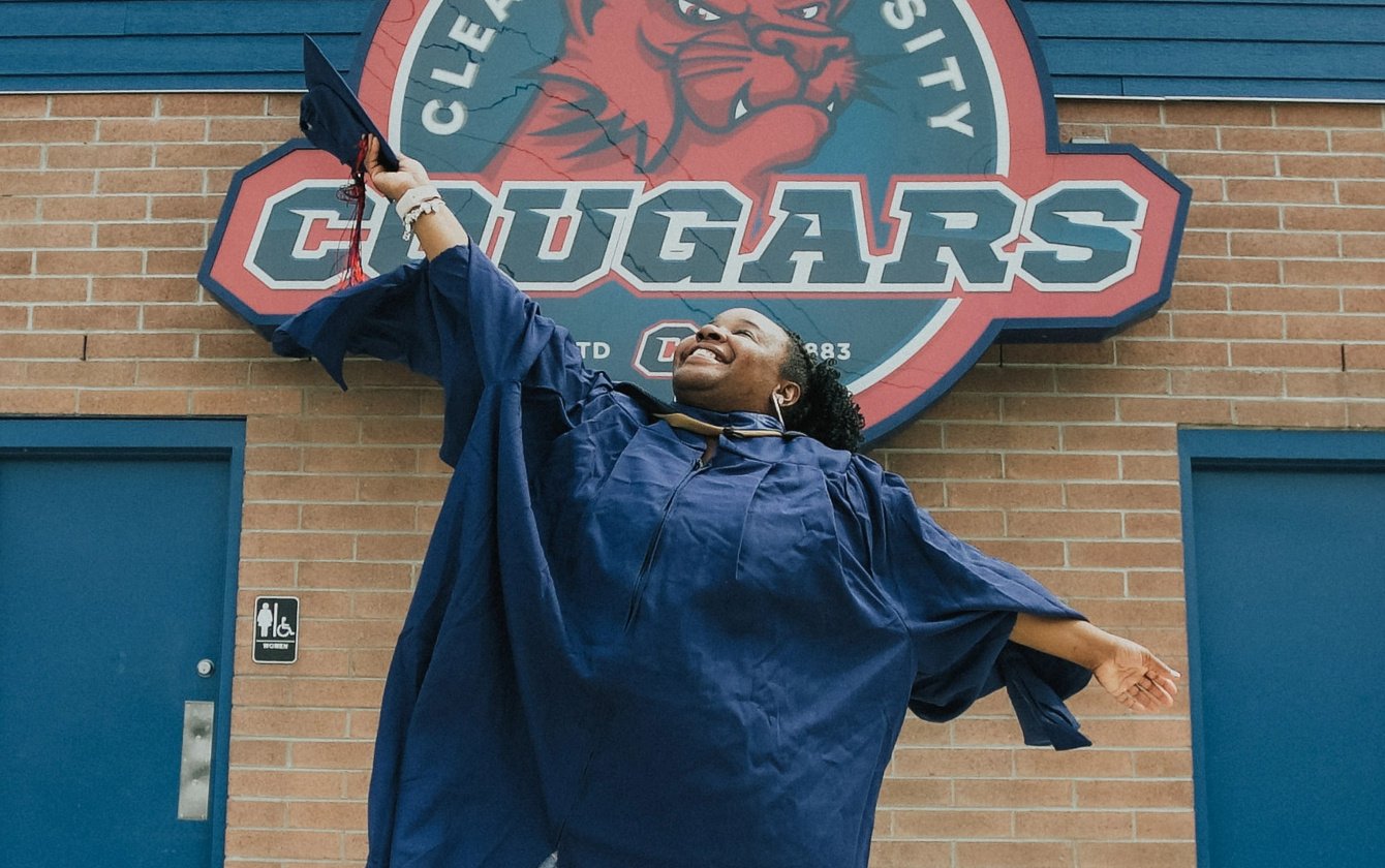 LaToya, a 2019 Lake Trust Foundation scholarship recipient, throws a graduation cap in the air in front of a Cleary University Cougars sign