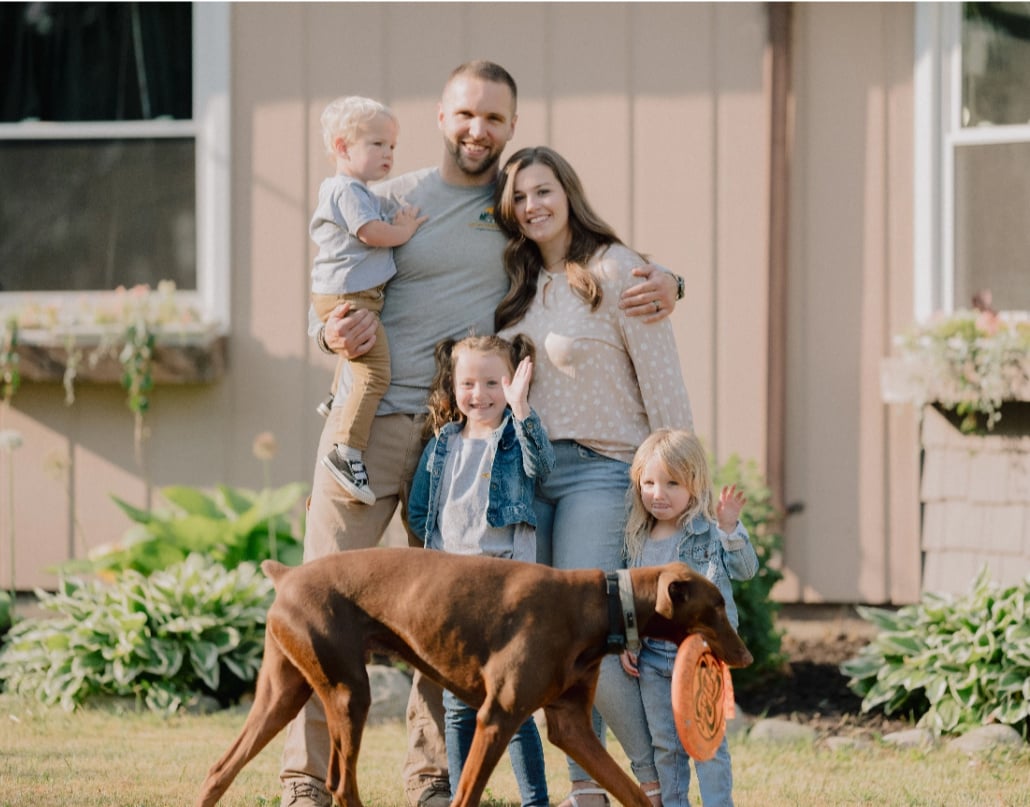 Colton, the owner of Hometown Plumbing, smiles with his wife, three children, and a brown dog with a frisbee in its mouth