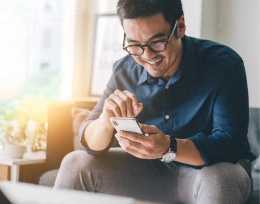 A man in glasses sits on a couch and looks at a phone