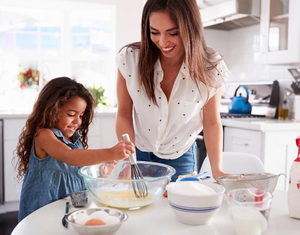 A woman in a kitchen teaches a young girl how to whisk eggs in a mixing bowl