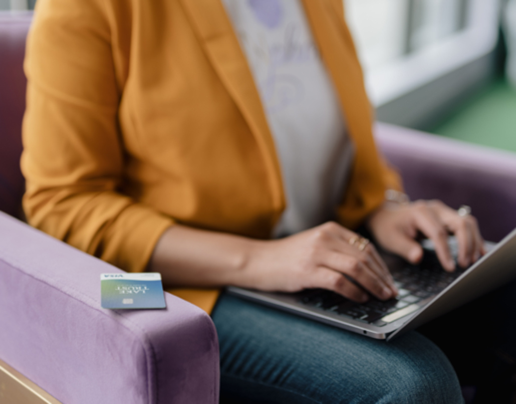 A woman in a yellow suit jacket sits on a purple chair with a laptop in her lap and a Lake Trust business debit card next to her