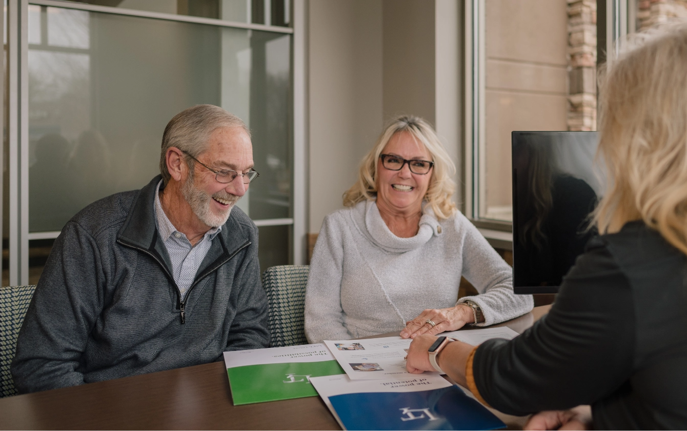 An older couple smile and laugh at a desk in a Lake Trust branch as a team member points to a piece of paper