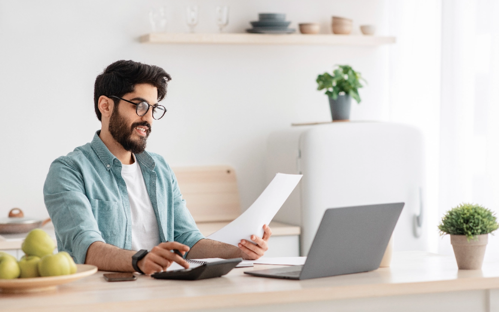 A man with a beard and glasses sits at a table and looks at a piece of paper while typing in a calculator