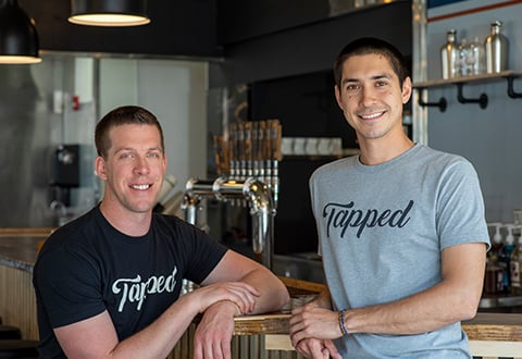 Kevin and Nick, the owners of Tapped Coffee, smile and pose in front of a row of beverage taps