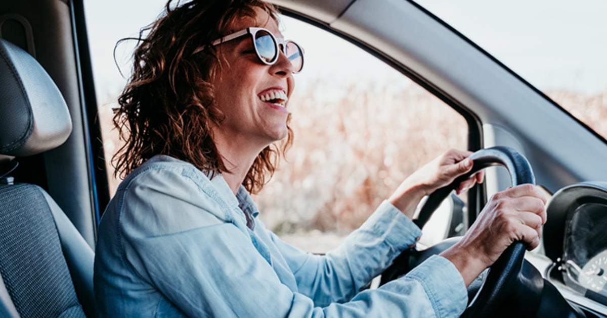 A woman in sunglasses sits in a car with her hands on the steering wheel