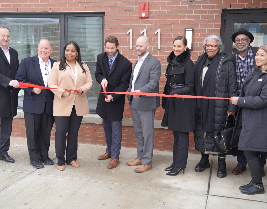 Nine people stand in a line as the person in the center cuts a red ribbon with scissors