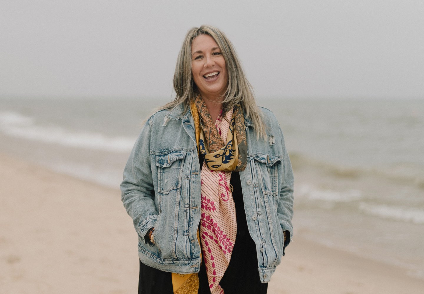 Nicole, the owner of More Love Collective, wears a jean jacket and scarf and smiles at a beach in Grand Haven