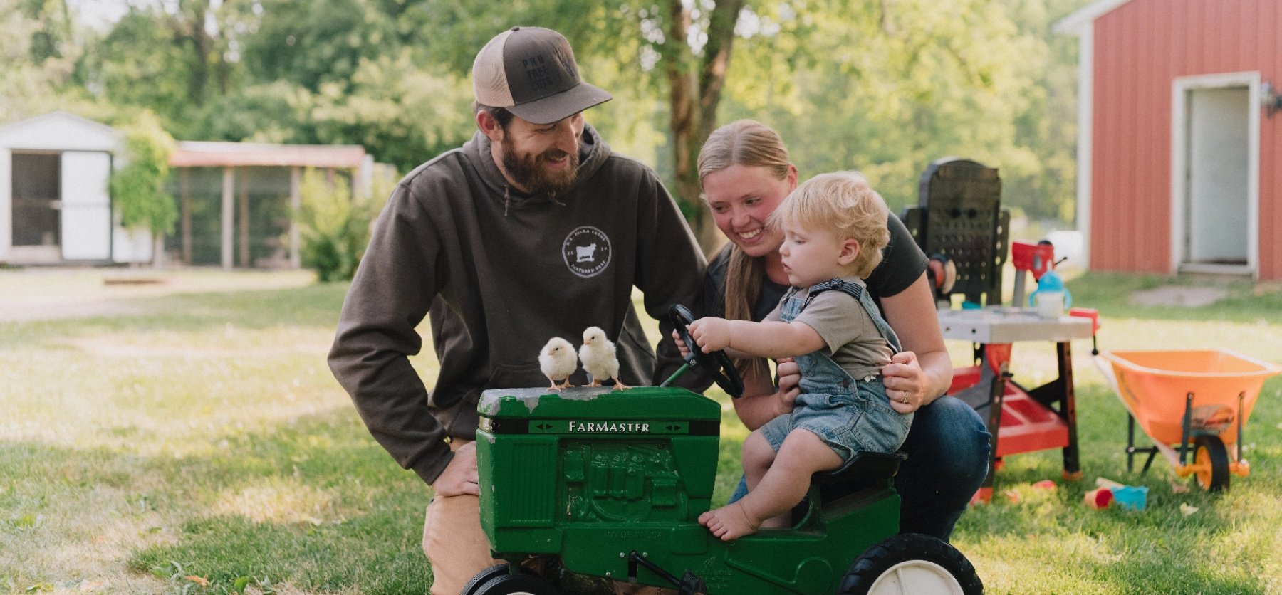 A young boy sits on a toy tractor by his parents